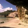 Una spiaggia sabbiosa riparata da una scogliera con vegetazione e un ponte in lontananza.