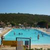 Piscina affollata con bagnanti, lettini e ombrelloni di paglia, colline verdi sullo sfondo.