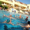 Persone in piscina che giocano, ombrelloni e edificio giallo sullo sfondo.