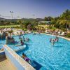 Piscina all'aperto affollata con giochi d'acqua, circondata da lettini e vegetazione.