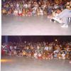 Un pubblico seduto all'aperto, composto da adulti e bambini, osserva qualcosa fuori campo.