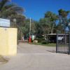 Ingresso di un campeggio con cancello aperto e cartello, circondato da alberi e palme.