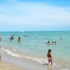 Spiaggia affollata con persone nel mare e sulla riva, sotto un cielo sereno.