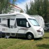 Camper bianco parcheggiato su erba con sedie pieghevoli.