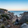 Uomo seduto su una roccia, guarda il mare tra scogli.
