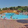Piscina all'aperto con molte persone che nuotano e giocano, un ponte di legno al centro.
