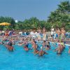 Persone giocano a pallavolo in piscina con ombrelloni gialli e palme sullo sfondo.