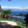 Vista su un mare blu con scogli, prato verde, panchine e pergola fiorita.