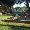Bambini che giocano in una sabbiera in un parco giochi con alberi e attrezzature colorate.