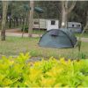 Una tenda verde in un campeggio con roulotte e cespugli gialli davanti.