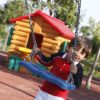 Un bambino su un'altalena blu in un parco giochi con una casetta di legno sullo sfondo.