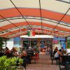 Area esterna di un bar con copertura e sedie nere. Persone sedute ai tavoli e bandiere appese.