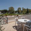 Terrazza con tavolo e sedie bianche, vista su un giardino con alberi e ombrelloni.