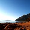 Costa rocciosa con terra rossa, mare calmo e montagne in lontananza sotto un cielo azzurro.