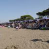 Una spiaggia con lettini e ombrelloni viola, persone rilassate e alberi sullo sfondo.