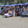 Persone sdraiate su lettini sotto ombrelloni blu in spiaggia, con un bambino che gioca.
