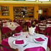 Ristorante con tavoli apparecchiati, tovaglie bordeaux e bottiglie in esposizione.