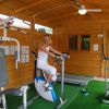 Donna su cyclette in una palestra di legno con attrezzi fitness e tappeto verde.