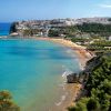 Spiaggia panoramica con mare azzurro e città collinare sullo sfondo.